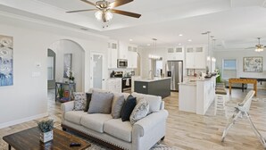 A spacious living room with a sofa, coffee table, and a ceiling fan. A kitchen with white cabinets and a dining area with a bench are visible in the background.