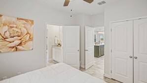 A bedroom with a large bed, white walls, and a ceiling fan. There is a bathroom visible through an open door, and a kitchen area in the background.