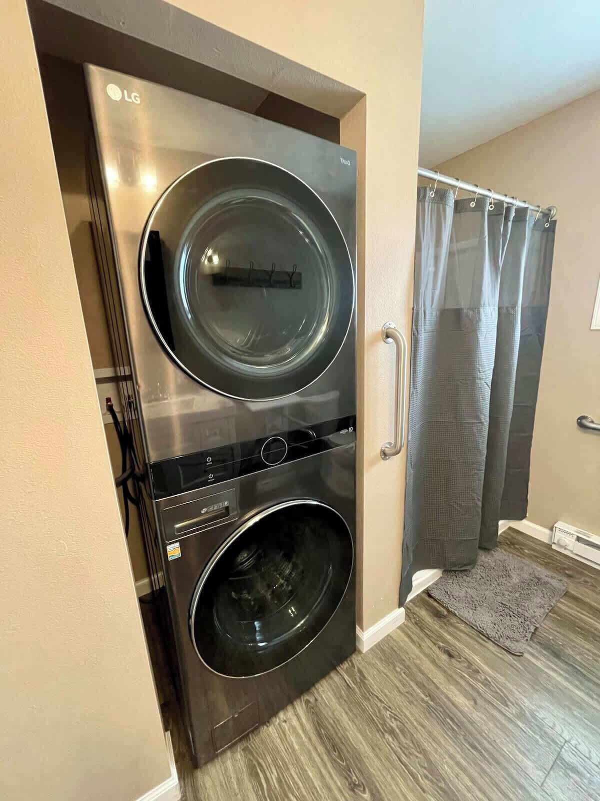 Een kleine, moderne wasruimte met een was- en droogcombinatie met dubbele deuren, een grijze douchegordijn en een houten vloer.
