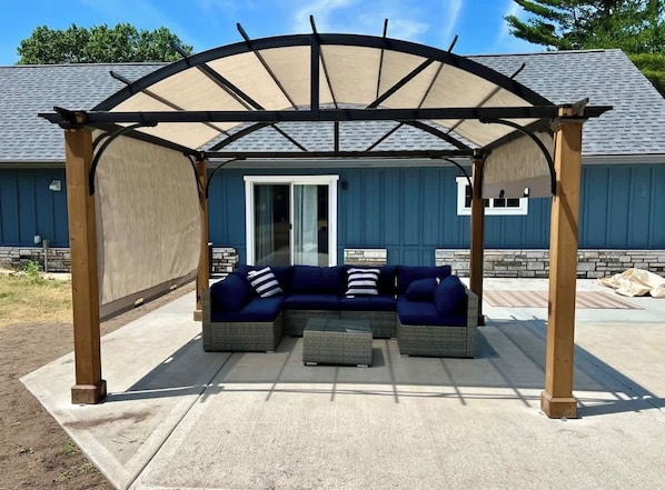 A covered outdoor seating area with a sectional sofa and ottoman, a pergola overhead, and a building with a blue exterior in the background.