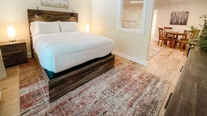 Un dormitorio con una cama grande, una mesita de noche, una lámpara de noche, una ventana con persianas, una zona de comedor con mesa y sillas, y una planta en maceta.