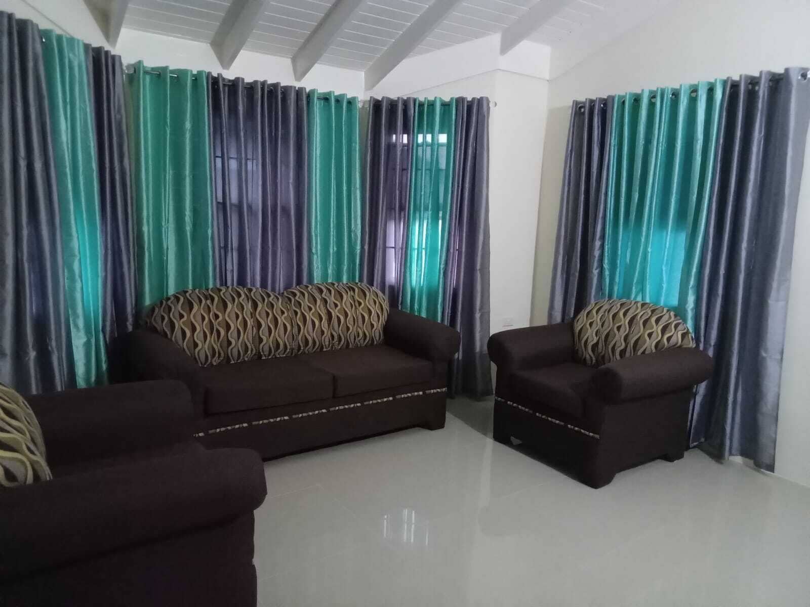 A living room with two sofas and a coffee table, large windows with curtains, and a high ceiling with exposed beams.