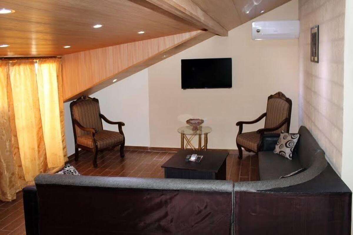 A living room with a dark brown sofa, two wooden armchairs, a small round table, and a flat-screen TV mounted on the wall.