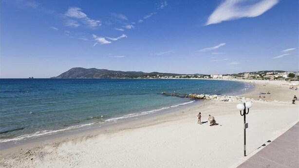 Une plage avec une eau d’un bleu limpide, un rivage sablonneux et quelques personnes. On aperçoit des bâtiments à l’arrière-plan et un lampadaire sur la droite.