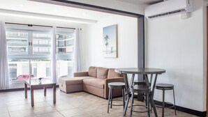 Een moderne woonkamer met een beige bank, een eettafel met glazen blad en krukken, een groot raam met gordijnen en een wandairconditioner.
