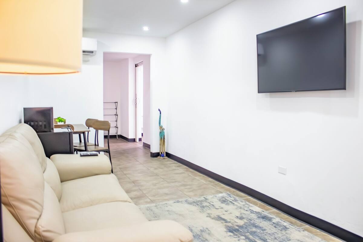 Un salon moderne avec un canapé beige, un téléviseur à écran plat fixé au mur, un coin repas avec une table et des chaises, et un couloir menant à une autre pièce.