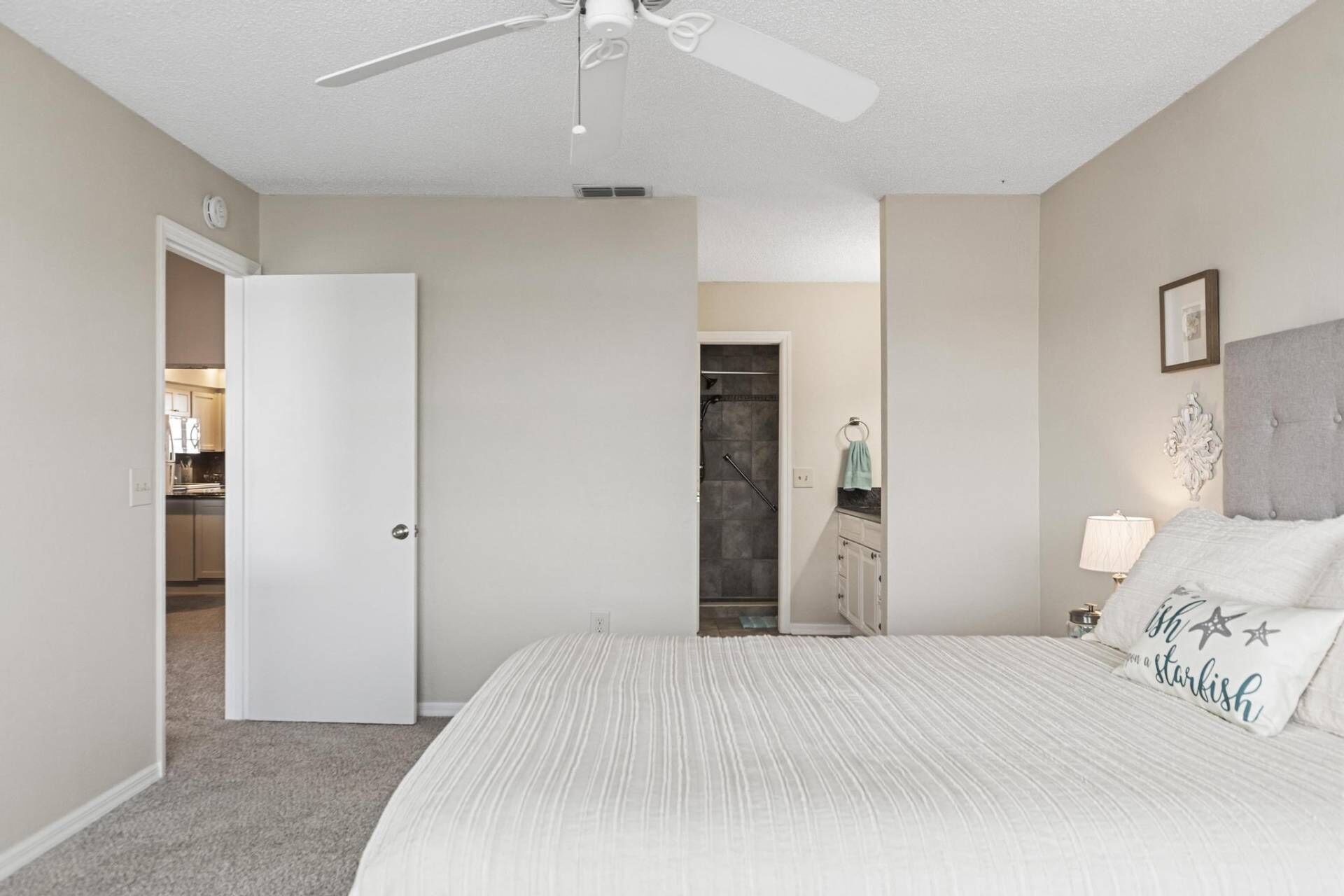 A bedroom with a bed, a ceiling fan, a nightstand with a lamp, and a bathroom visible through an open door.