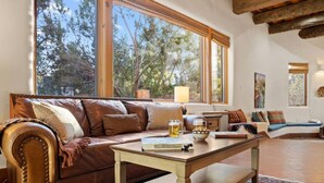 A cozy living room with a leather sofa, a coffee table, and a view of trees through large windows. - Upscale Home In North Hills (Santa Fe)