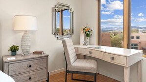 Ein modernes Schlafzimmer mit Schreibtisch, Stuhl, Lampe, Kommode und einem großen Fenster mit Blick auf Gebäude und Berge.