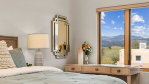 Ein Schlafzimmer mit Bett, Schminktisch, Spiegel, Blumen in einer Vase und einem Fenster mit Bergblick.