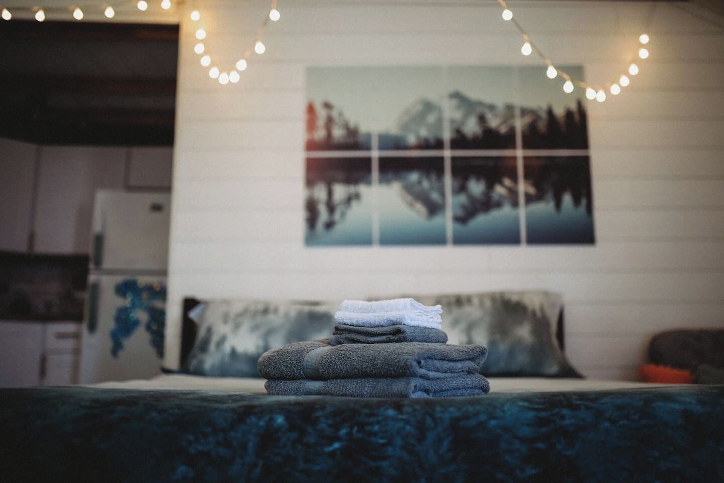 Una cama bien tendida con toallas dobladas, una pared con un cuadro enmarcado y luces de adorno.