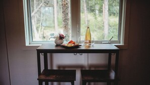 Ein kleiner Tisch mit einer Weinflasche, Gläsern und einem Teller Essen neben einem Fenster mit Blick auf Bäume.
