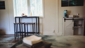 Ein gemütliches Zimmer mit Holzboden, einem kleinen Tisch mit zwei Stühlen, einem Fenster mit Vorhängen sowie einer Küchenzeile mit Kaffeemaschine und Ablagefläche.