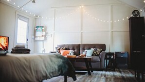 Ein gemütliches Wohnzimmer mit einer Couch, einem Couchtisch und einem Fernseher. An der Decke befinden sich Lichterketten und ein Stehlüfter.