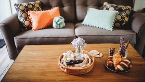 Ein gemütliches Wohnzimmer mit einem braunen Sofa, das mit gemusterten Kissen geschmückt ist, einem Holztisch mit einem gewebten Korb, einer Blumenvase und einer Schüssel Croissants.
