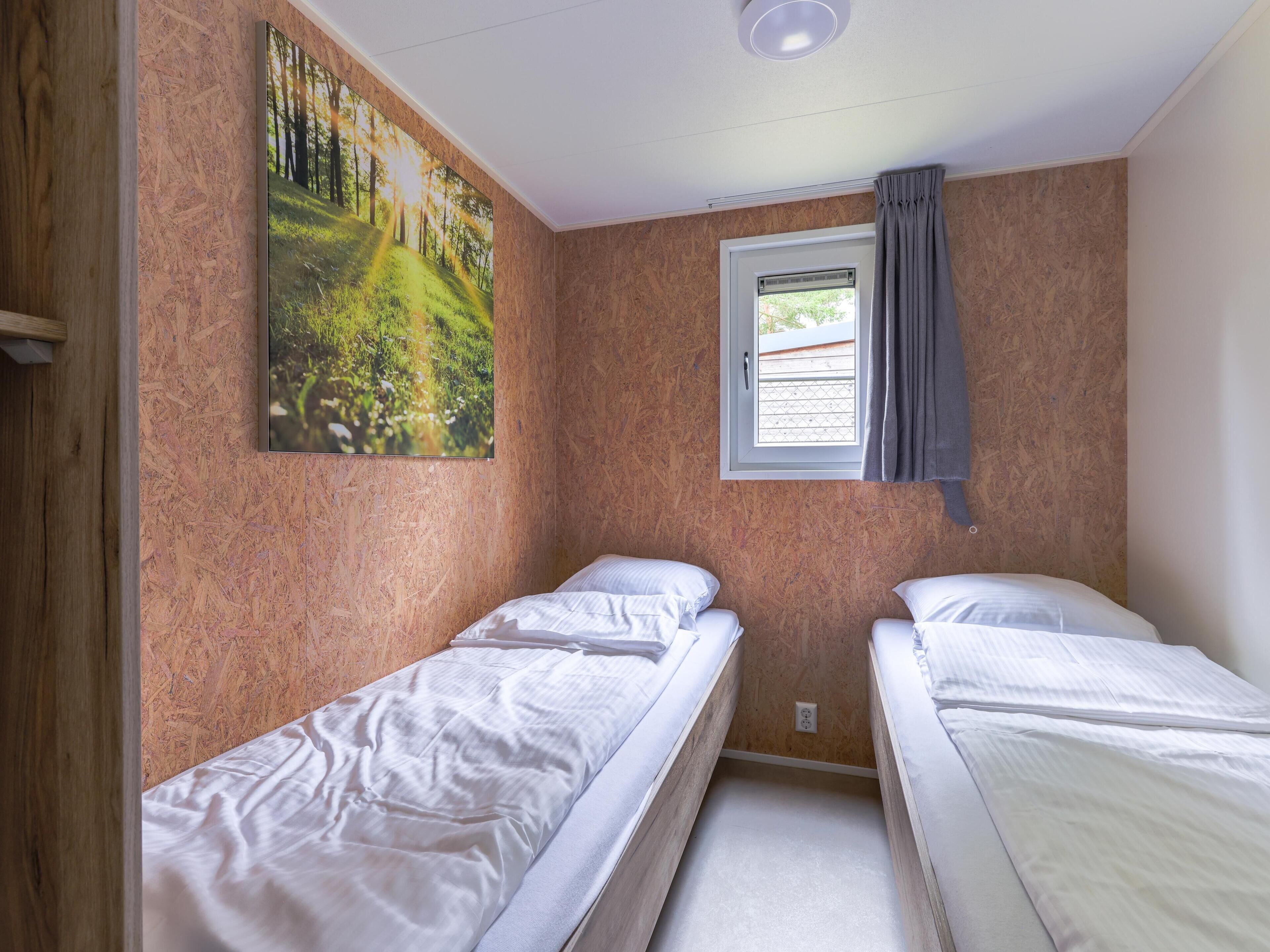 Ein Zimmer mit zwei Einzelbetten, einem fensterbelaubten Fenster, einem Waldgemälde und einer Deckenleuchte.