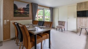 A dining area with a table set for four, a television mounted on the wall, and a window with curtains. - chalet with covered terrace on campsite with pool (Vessem)