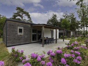 A modern wooden cabin with a patio, surrounded by lush greenery and purple flowers.