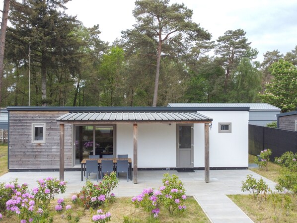 A modern single-story house with a covered patio, a dining area with chairs, and a small garden with pink flowers.
