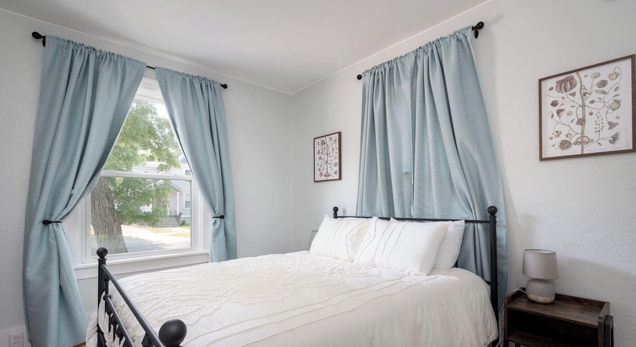 Un dormitorio con cama, cortinas, mesita de noche, lámpara y un cuadro en la pared.