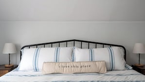 Una cama con estructura metálica negra, ropa de cama blanca con rayas azules y una almohada con la frase 'I love this place'.