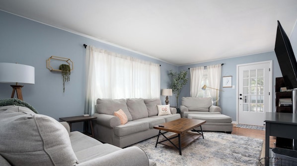 A modern living room with a grey sofa, a wooden coffee table, and a large window with white curtains. - Holland Hideaway (Holland)