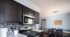 A modern kitchen with dark cabinets, a stainless steel microwave, a gas stove, and a tiled backsplash. There is a wooden dining table with chairs and a framed map on the wall.