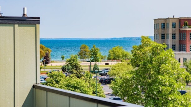 A view of a parking lot with cars and trees, a building with a balcony, and a body of water in the background. - Rooftop Condo in TC w/ Bay Views (Traverse City)