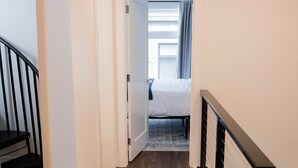 A hallway with wooden flooring, a staircase with black railings, and a bedroom with a bed and curtains. - Rooftop Condo in TC w/ Bay Views (Traverse City)