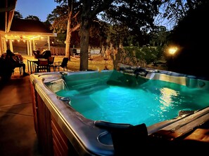 A hot tub with a wooden deck and chairs, surrounded by trees and a covered area with string lights. - Picayune Retreat: Spa/EV/Play-structure/Pet-loving/Games and more'' (Coarsegold)