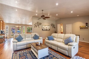 A spacious living room with two white leather sofas, a wooden coffee table, and a ceiling fan. - Picayune Retreat: Spa/EV/Play-structure/Pet-loving/Games and more'' (Coarsegold)