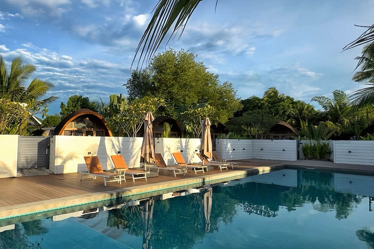 Piscina con sillones de descanso y sombrillas, rodeada de vegetación y paredes blancas con estructuras arqueadas.