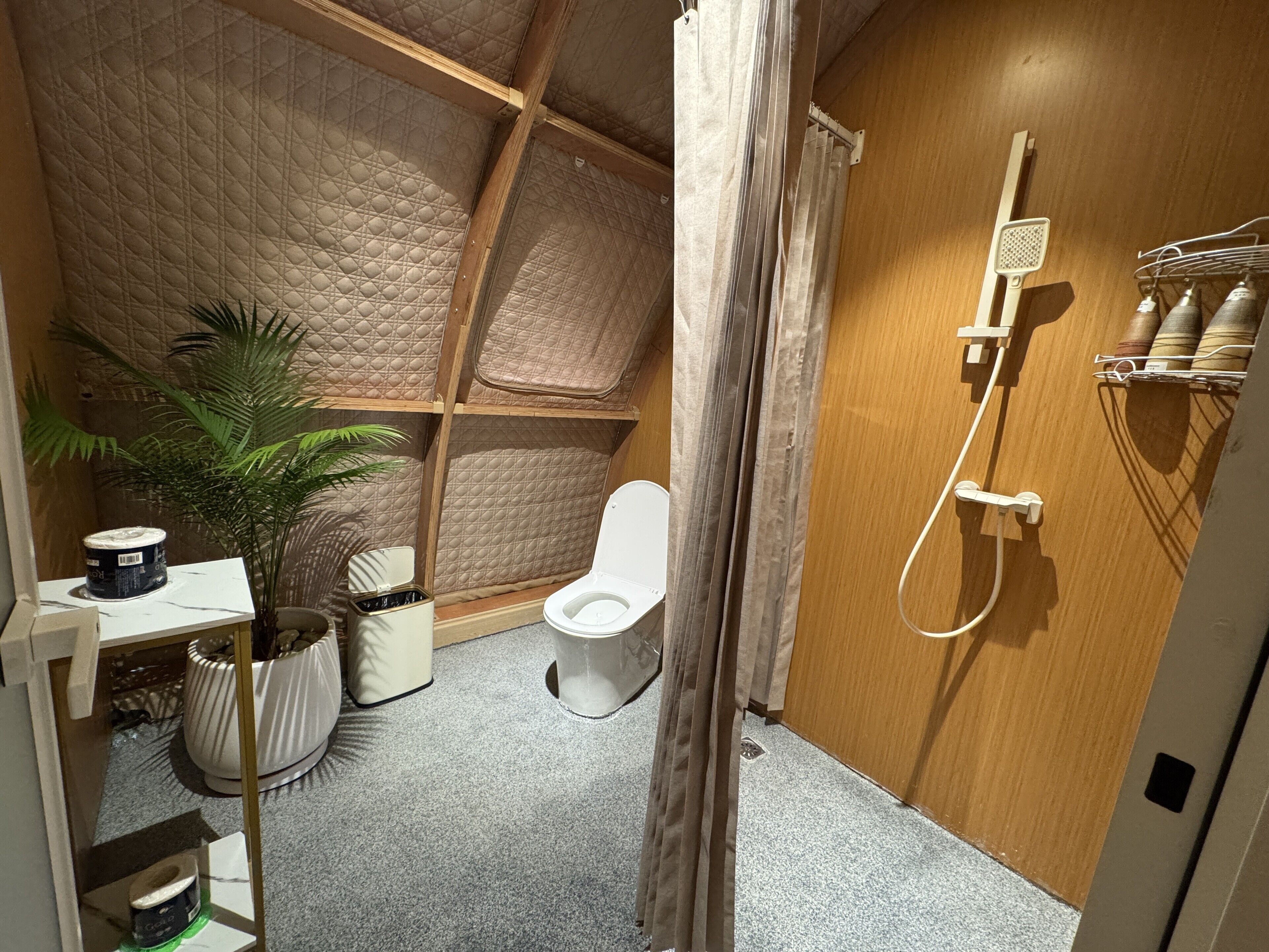 A compact bathroom with a wooden ceiling, a white toilet, a shower with a handheld showerhead, and a small shelf with toiletries.