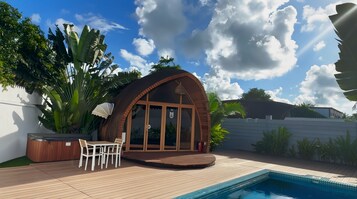 Un área de piscina exterior moderna con un deck de madera, un espacio de estar cubierto y exuberante vegetación tropical.