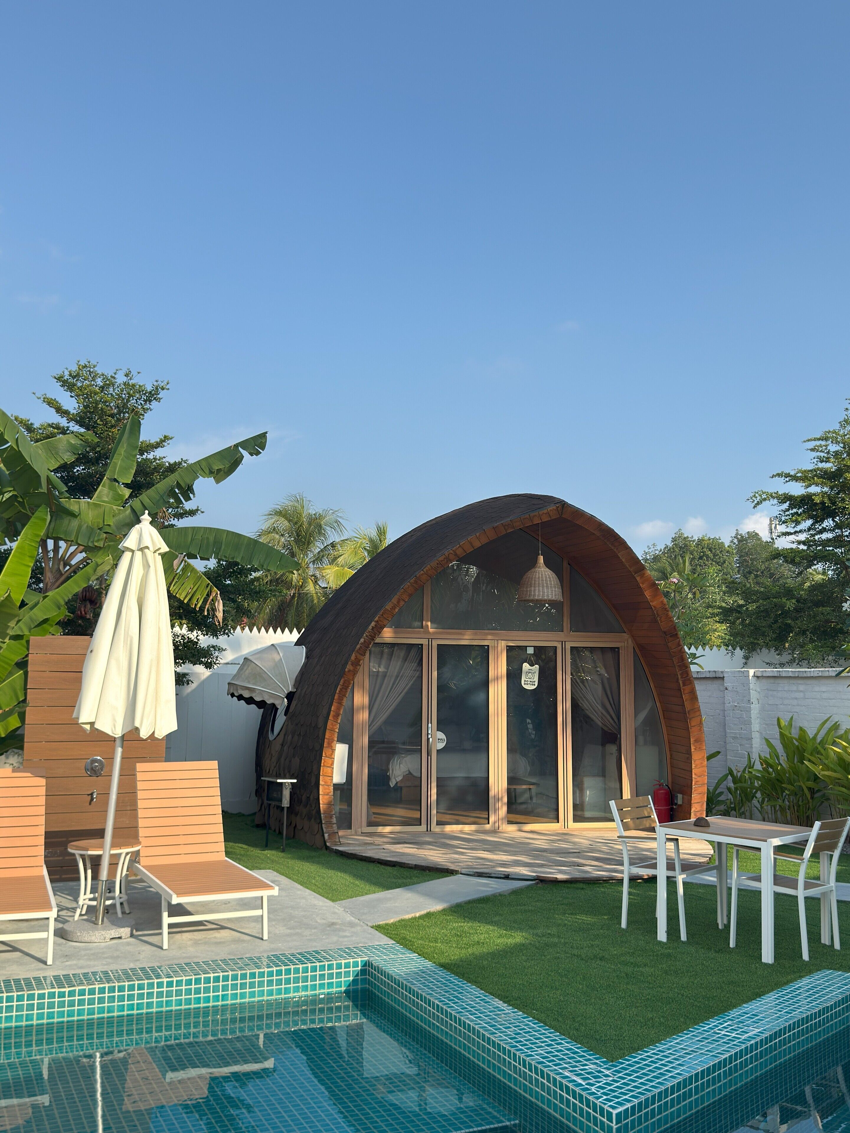 A modern, dome-shaped building with a glass door and windows, surrounded by a swimming pool and outdoor furniture.