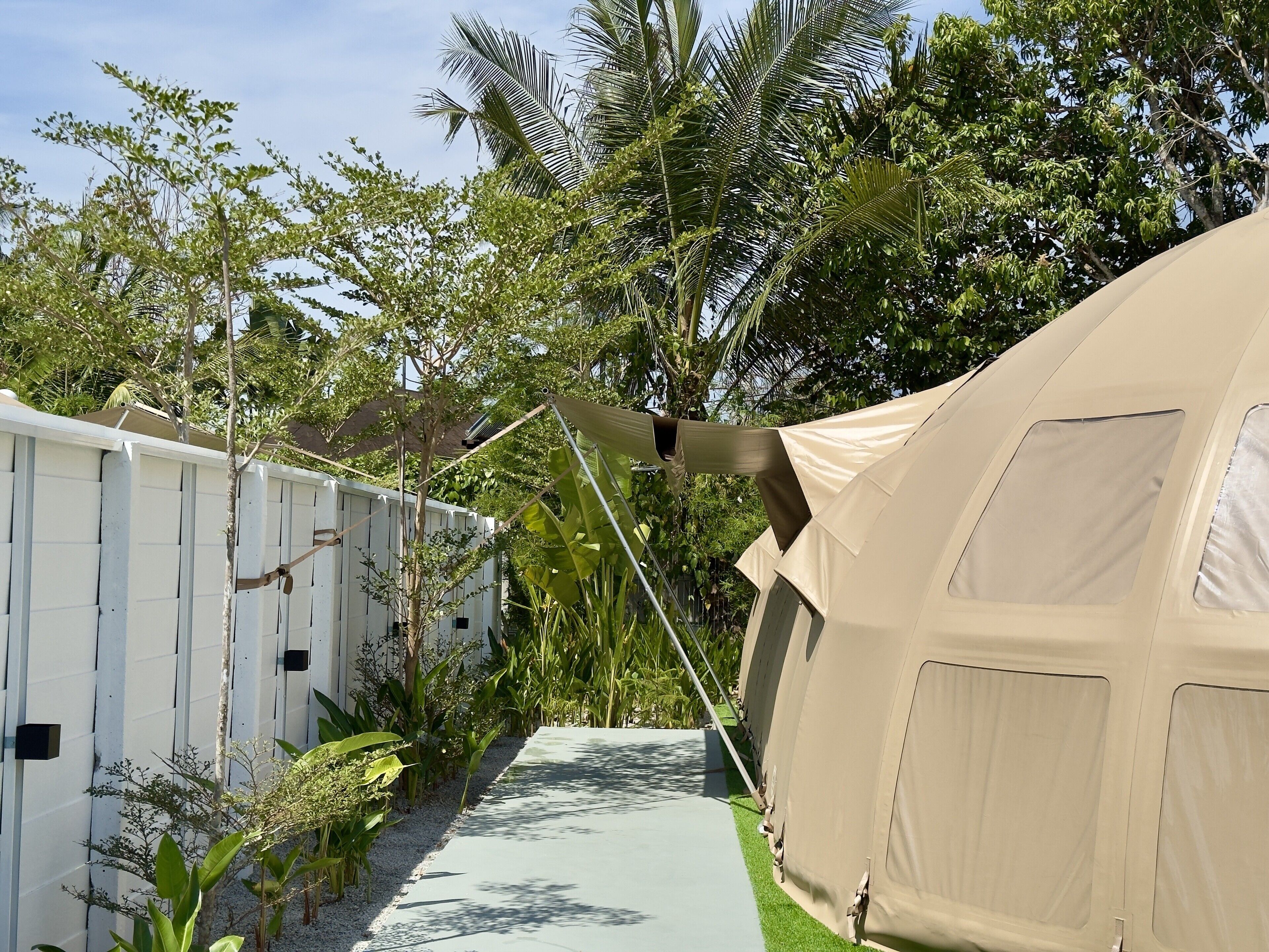 A tented accommodation area with a pathway leading to a white building, surrounded by lush greenery.