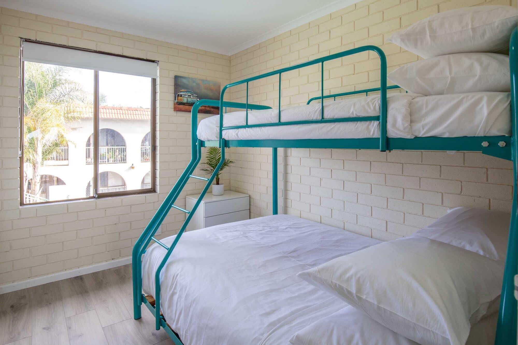 Een studentenkamer met een stapelbed, een raam, een klein nachtkastje en een bloempot.