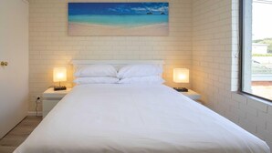 Une chambre d’hôtel avec un lit, des murs blancs, un tableau représentant une plage, deux lampes de chevet et une fenêtre.