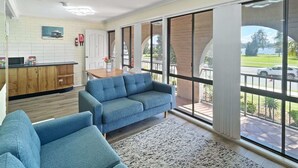 Un salon moderne avec un canapé et des fauteuils, un coin repas avec une table et des chaises, et de grandes fenêtres offrant une vue sur l’extérieur.