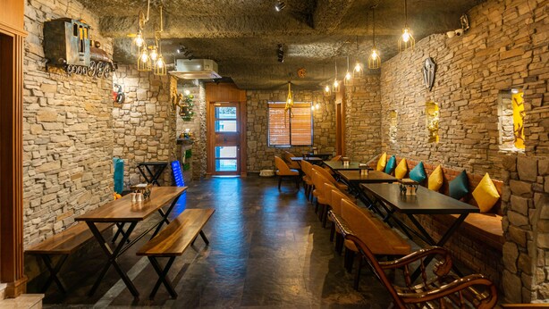 Een restaurant met stenen muren, houten tafels en banken. Er hangen lampen en een deur die naar buiten leidt.