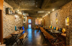 A restaurant with stone walls, wooden tables, and benches. There are hanging lights and a door leading outside. - Hotel Gems Inn (Vikasnagar)