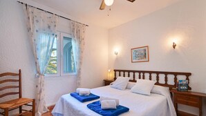 Habitació d'hotel amb llit, dos coixins, una taula de nit amb un llibre, una cadira, una finestra amb cortines i un lluminari fixat a la paret.