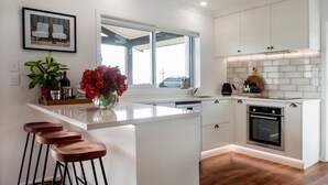 A modern kitchen with white cabinetry, a large window, and a bar area with stools. - Poolside Stay with City Views | Close to the Redwoods for Forest Adventures (Rotorua)