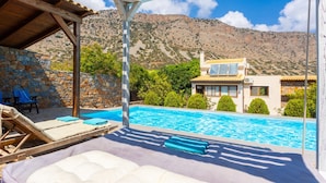 A pool area with lounge chairs and a pergola, a stone wall, and a view of a mountain and a modern house. - Villa Elounda Plaka in Plaka (Agios Nikolaos)