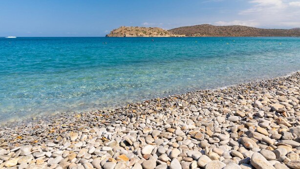 A pebbled beach with clear turquoise water and distant rocky hills. - Villa Elounda Plaka in Plaka (Agios Nikolaos)