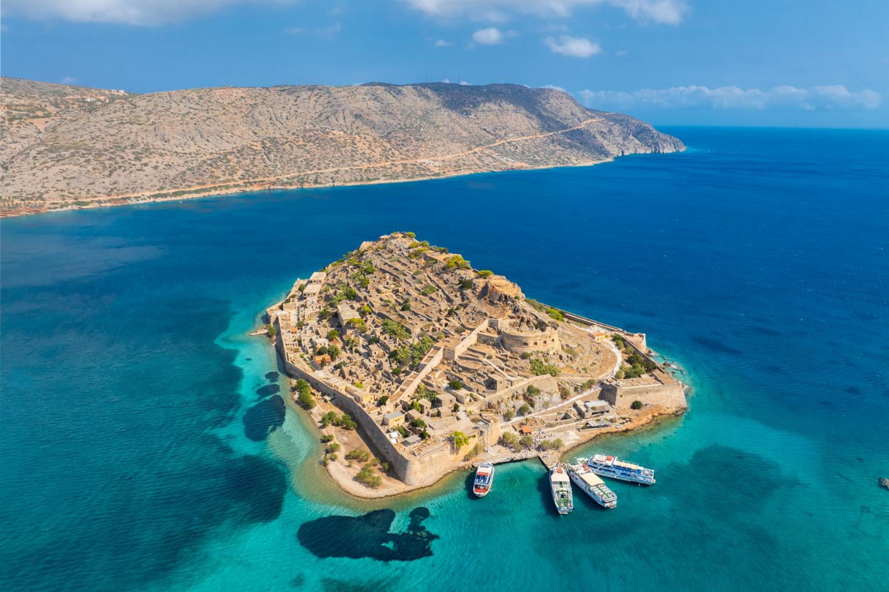 Vista aérea de una isla costera con una estructura fortificada, aguas azules cristalinas y varios barcos amarrados cerca de la orilla.