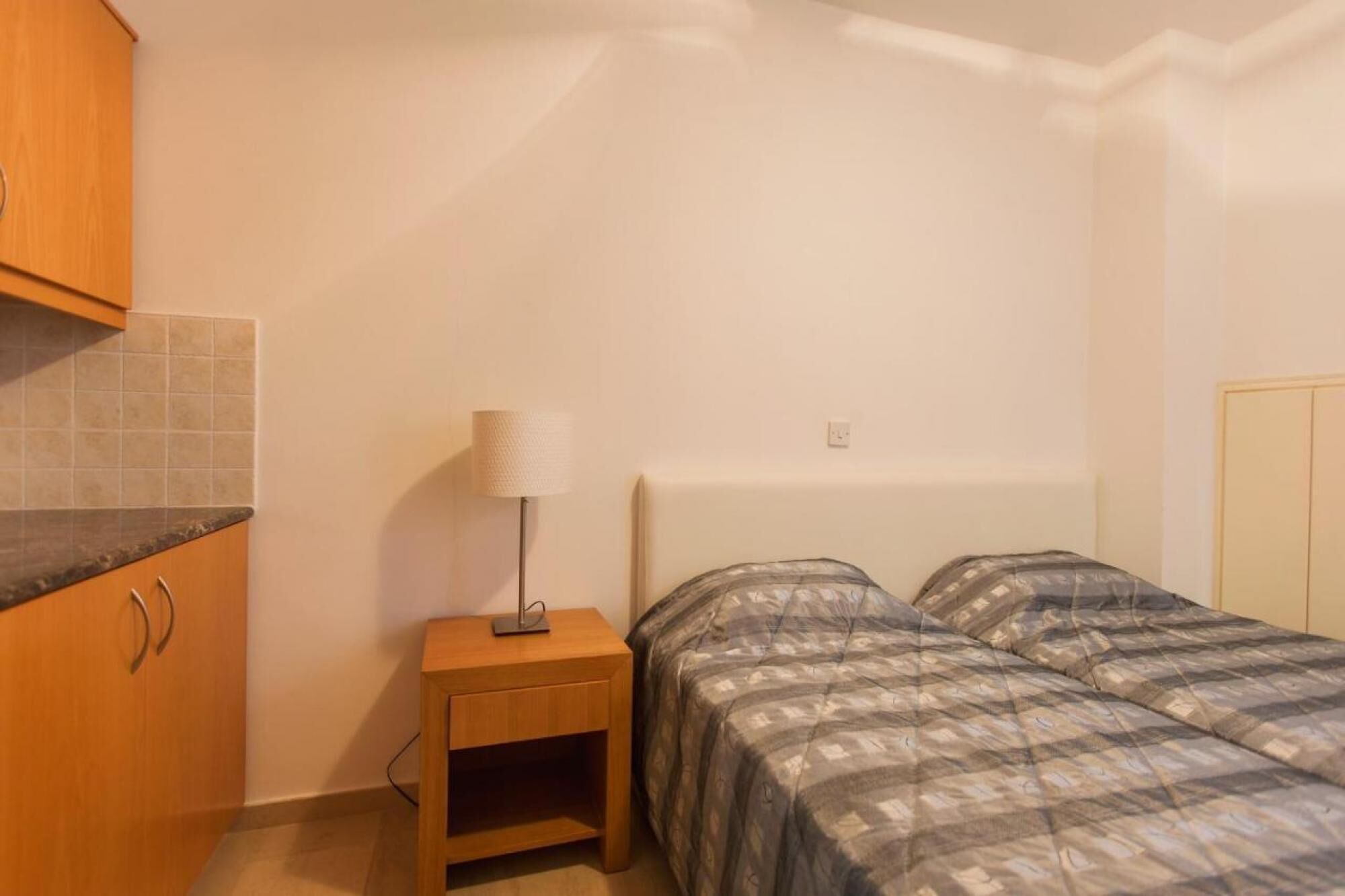Une petite pièce bien éclairée, avec un lit, une table de chevet et une armoire en bois.