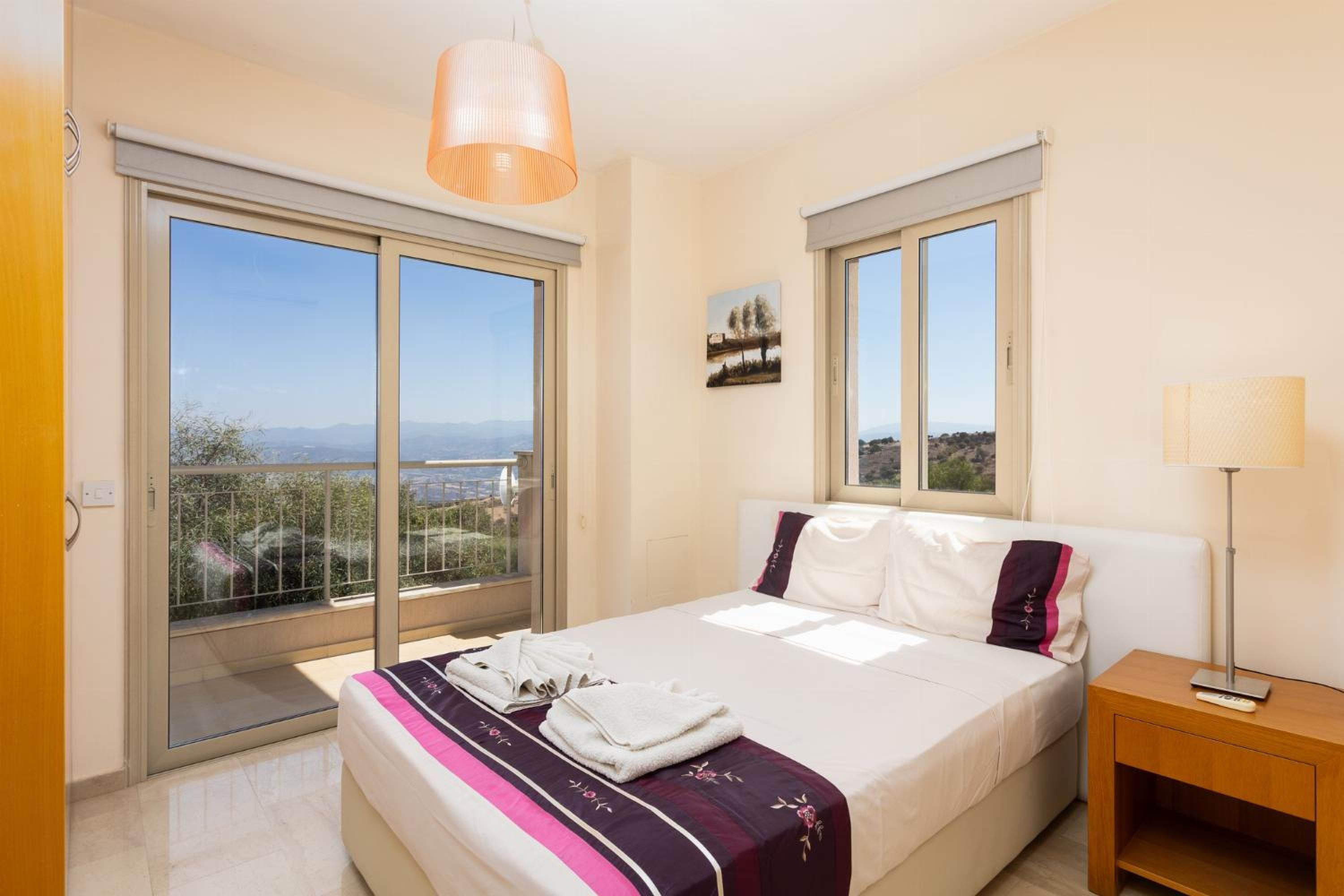 A bedroom with a bed, a nightstand, a lamp, and a view of the outdoors through large windows.