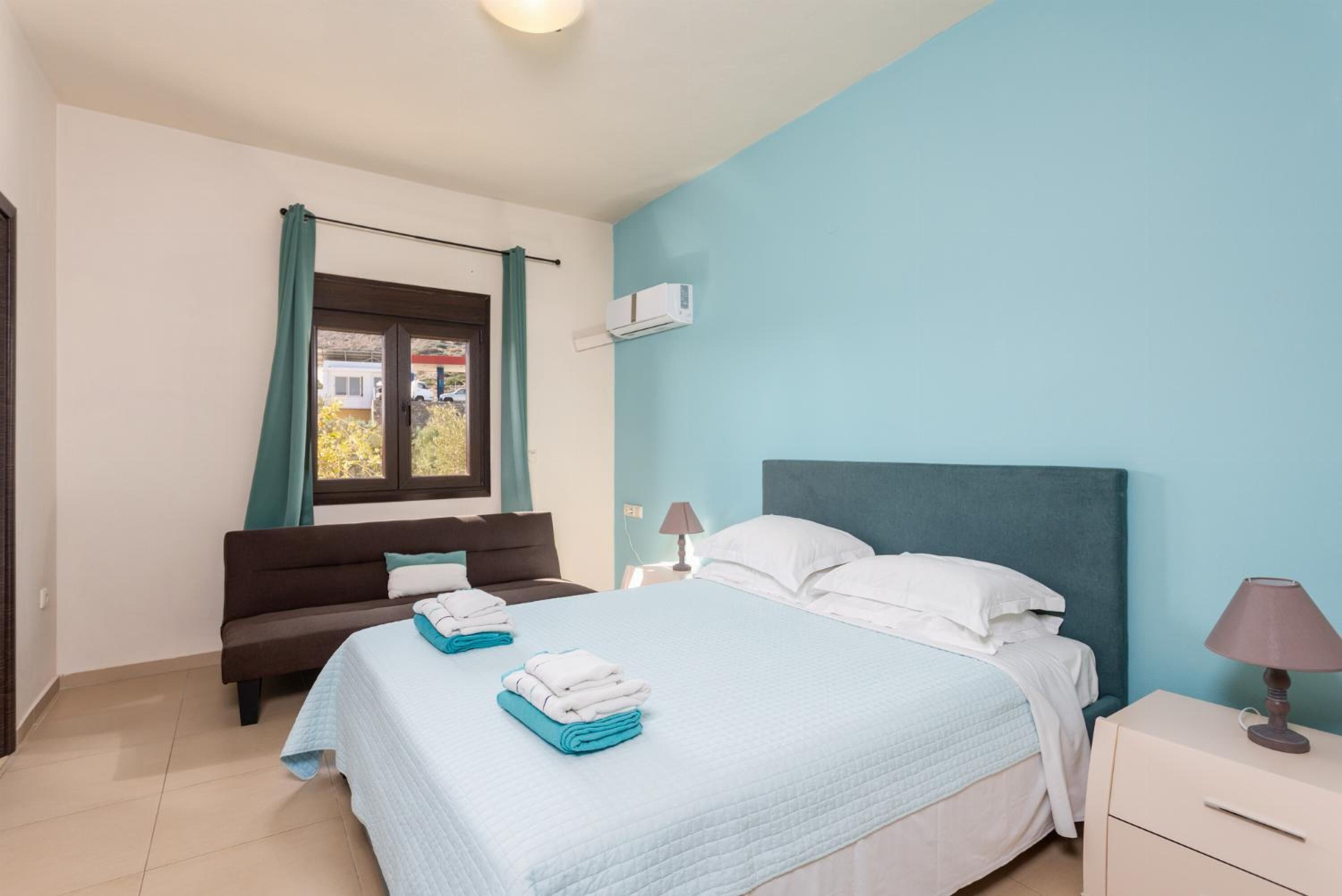 Une chambre à coucher avec un lit, une table de chevet, une lampe, une fenêtre avec des rideaux et une vue sur l’extérieur.