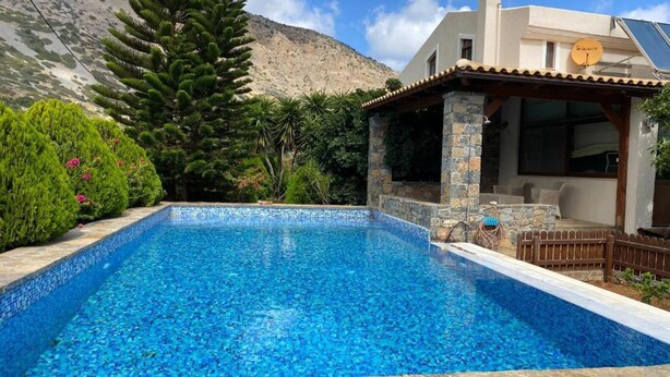 A rectangular swimming pool with clear blue water, surrounded by lush greenery and a stone wall. - Villa Elounda Blue in Plaka (Agios Nikolaos)